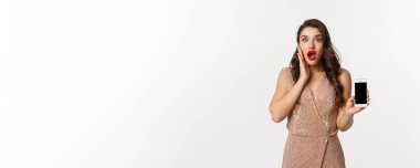 İnternetten alışveriş. Lüks elbiseli zarif bir kadın akıllı telefon ekranı gösteriyor, kameraya hayran bakıyor, beyaz arka plan.