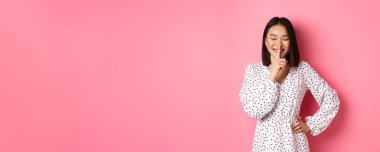 Moda elbiseli güzel bir Koreli kadın. Sır tutmak istiyor. Yumuşak gülüşü ve kapalı gözleriyle pembe arka planda duruyor..