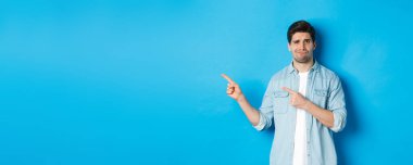 Şüpheci yetişkin adam parmaklarını sol tarafı işaret ediyor, şüpheli ve kararsız görünüyor, mavi arka planda duruyor..