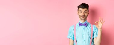 Papyon ve pantolon askısı takmış gülümseyen beyaz adam iyi bir jest sergiliyor, havalı bir şey öneriyor, pembe arka plandaki tanıtıma övgüler yağdırıyor..