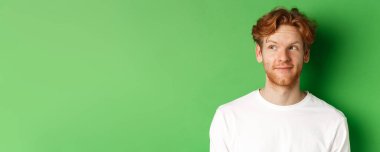 Duygular ve moda konsepti. Yakışıklı kızıl saçlı, sakallı, dağınık saçlı, sola bakan, gülümseyen, yeşil arka planlı erkek manken..