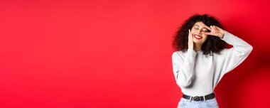 Güzellik. Kıvırcık saçlı, kırmızı dudaklı, makyajlı, yüzünde V işareti olan, gamsız gülümseyen, stüdyo arkasında duran güzel bir bayan..