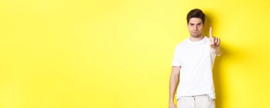 Ciddi bir adam hoşnutsuz, bir şeyi yasaklamak için parmağını sallıyor, seni uyarıyor, sarı arka planda duruyor..