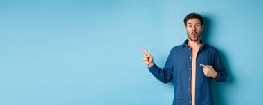 Şaşırmış ve şaşırmış beyaz adam hayranlıkla soluk soluğa kalmış, sol logoyu işaret ediyor, müthiş bir tanıtım yapıyor, mavi arka planda duruyor..