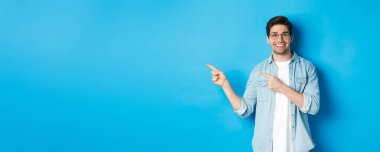 Gözlüklü genç yakışıklı adam reklamını yapıyor, gülümsüyor ve parmaklarını sola gösteriyor, duyuru yapıyor, mavi arka planda duruyor..