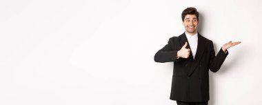 Siyah takım elbiseli yakışıklı bir iş adamı. Başparmak kaldırıyor ve ürününüzü beyaz fotokopi alanının üzerinde tutuyor. Beyaz arka planda duruyor..