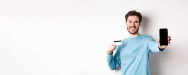 E-ticaret ve alışveriş konsepti. Gülümseyen beyaz adam plastik kredi kartı ve boş akıllı telefon ekranı gösteriyor. Çevrimiçi uygulama öneriyor..