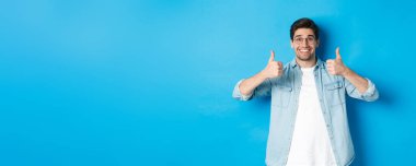 Gözlüklü ve gündelik giysiler giyen çekici bir genç. Onaylama için başparmak gösteriyor. Bir şey gibi..