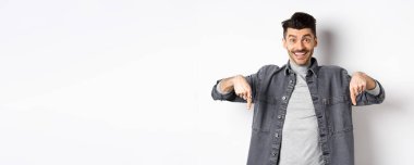 Heyecanlı, gülümseyen erkek manken parmaklarını aşağıya doğru gösteriyor, mutlu yüzlü logoyu gösteriyor, havalı bir promosyon sergiliyor, beyaz arka planda duruyor..