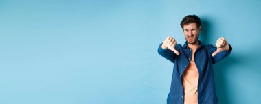 Tiksinç adam olumsuz görüş bildiriyor, baş parmaklarını aşağı ve dillerini gösteriyor, üzgün, kötü bir şeyi onaylamıyor, mavi arka planda duruyor..