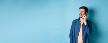 Yakışıklı beyaz adam cep telefonuyla konuşuyor, boş yere bakıyor kaygısız bir gülümsemeyle, mavi arka planda duruyor..