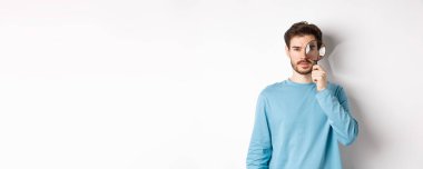 Adam bir şey arıyor, büyüteçten kameraya bakıyor, beyaz arka planda duruyor..