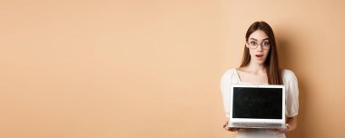 Gözlüklü, hayret verici yüzü olan, bej arka planda duran dizüstü bilgisayarlı genç bir kadın..