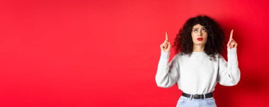 Endişeli, kıvırcık saçlı, kaşlarını çatmış ve şüpheli görünen, tereddütlü ya da endişeli yüzüyle kırmızı arka planda duran genç bir kadın..