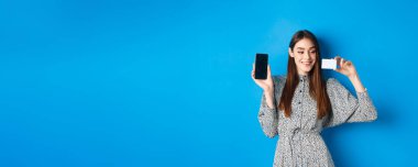 İnternetten alışveriş. Güzel bir kadın model boş cep telefonu ekranı ve plastik kredi kartı gösteriyor, gülümsüyor, internet mağazasından alıyor, mavi arka plan..