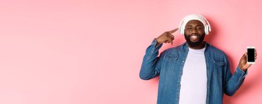Mutlu zenci adam müzik dinliyor, kulaklıkları işaret ediyor ve gülümsüyor, cep telefonu ekran uygulamasını veya çalma listesini gösteriyor, pembe arka planda duruyor..