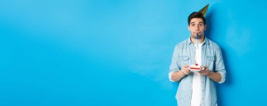 Doğum günü pastası tutan yakışıklı genç adam, parti şapkası takıyor ve düdük çalıyor, mavi arka planda duruyor..