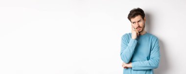 Tişörtlü sakallı endişeli genç adam, dalgın dalgın bakıyor ve düşünüyor, beyaz arka plana karşı sorunlu duruyor. Boşluğu kopyala