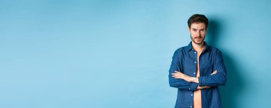 Kendine güveni tam genç bir adam kendini profesyonel hissediyor, kollarını göğsünde kavuşturuyor ve sırıtıyor, mavi arka planda duruyor..