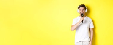 Kulaklıklı bir adam mikrofon tutuyor, karaoke şarkıları söylüyor, beyaz elbiseli sarı arka planda duruyor..