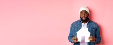 Emlak konsepti. Üzgün ve yorgun siyah adam kameraya gülmeden bakıyor, kağıt ev modelini tutuyor, pembe arka planda duruyor..