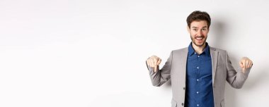 Hey, şuna bak. Takım elbiseli mutlu işadamı parmaklarını aşağı uzatıp gülümsüyor, etkinliğe davet ediyor, bağlantıyı gösteriyor, beyaz arka planda duruyor..