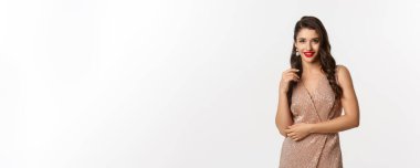 Kırmızı rujlu zarif bir kadın, şık küpeler memnun görünüyor, kış tatilini kutluyor, yeni yıl partisi için elbise giyiyor, beyaz arka planda duruyor..