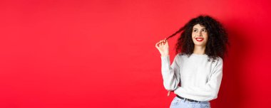 Güzel bir kadın model kıvırcık saçla oynuyor ve mutlu görünüyor, bir şey hakkında hayaller kuruyor, kırmızı arka planda duruyor..