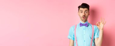 Papyonlu ve pantolon askısı takmış etkileyici bir adam hayret verici bir jest yapıyor, harika bir şey, pembe arka planda duruyor..