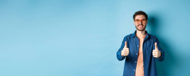 Gözlüklü mutlu genç adam başparmağını kaldırıyor, gözlükçü dükkanını tavsiye ediyor, mavi arka planda duruyor..