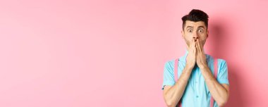 Şok olmuş bir adam bakıyor, elleriyle ağzını kapatıyor ve pembe arka planda durup kameraya bakıyor..