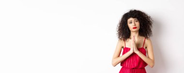 Kıvırcık saçlı kadın, elbise giyiyor ve yardım için yalvarıyor, el ele tutuşuyor, iyilik istiyor, beyaz arka planda duruyor..