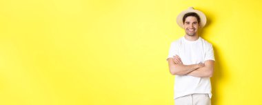 Turizm. Yakışıklı genç adam mutlu görünüyor, gezmek için hasır şapka giyiyor, göğsünde çapraz eller ve gülümsüyor, sarı arka planda duruyor..