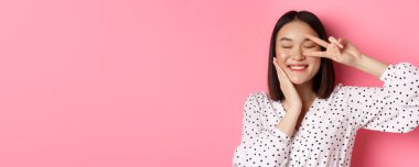 Güzellik ve yaşam tarzı konsepti. Güzel Asyalı bir kadının barış işareti gösterip yanaklarına dokunduğu, kameraya mutlu bir şekilde gülümsediği, pembe arka planda durduğu yakın plan..