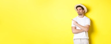 Turizm ve yaşam tarzı kavramı. Memnun olmayan erkek turist şikayetçi, sol üst köşedeki tanıtıma bakıyor ve işaret ediyor, hayal kırıklığıyla, sarı arka planda duruyor..