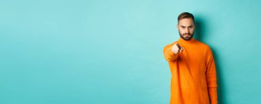 Kendine güvenen adam seni suçlar, kaşlarını çatar, kamerayı işaret eder, suçlar, yargılar, açık mavi arka planda durursun..