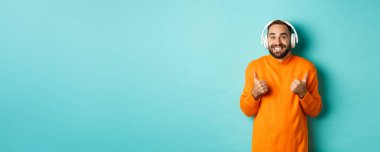 Kulaklıkla müzik dinleyen hayret verici yetişkin bir adam, kameraya bakıyor. Etkilenmiş ve onaylanmış. Tavsiye ediyor, turkuaz arka planda duruyor..