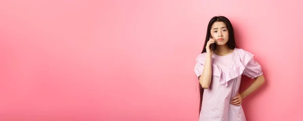 Sad asian girl calling someone, holding phone and talking, standing upset against pink background.