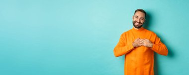 Minnettar adam gülümsüyor, kalbi tutuyor, teşekkür ediyor, hediyeyle duygulanmış hissediyor, turkuaz arka planda duruyor..