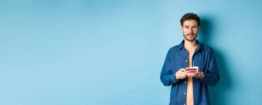 Doğum gününü kutlayan gülümseyen genç bir adam. Elinde mumla doğum günü pastası tutarken mavi arka planda dikilip kameraya bakarken..