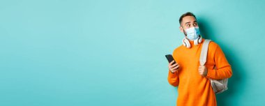 Yüz maskeli genç adam cep telefonu kullanıyor, sırt çantası tutuyor, şaşkın şaşkın bakıyor, açık mavi arka planda duruyor..