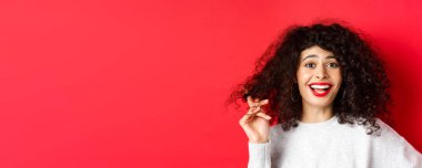 Kıvırcık saçlarıyla oynayan ve kırmızı arka planda duran komik bir şeye gülen mutlu kadının yakın çekimi..