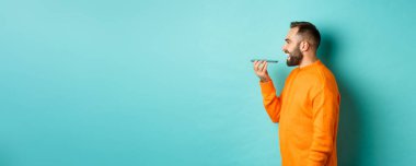 Yakışıklı sakallı bir adamın sesli mesaj kaydetme profili, cep telefonu tutmak ve hoparlörden konuşmak, mutlu bir şekilde gülümsemek, turkuaz arka planda durmak..