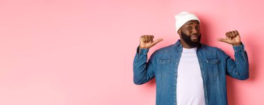 Kendinden emin ve küstah siyah adam kendini işaret ediyor, kameraya bakıyor kendinden emin, gösteriş meraklısı, pembe arka planda duruyor..