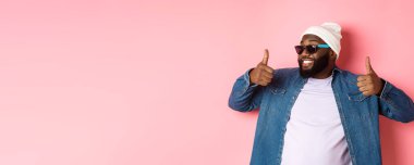 Güneş gözlüklü mutlu ve tatmin olmuş Afro-Amerikan adam. Başparmak kaldırıyor, övüyor ve promosyon gibi pembe arka planda dikiliyor..