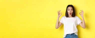 Etkilenmiş Koreli kız, tamam işaretleri gösteriyor ve şaşırmış görünüyor, sarı arka planda duruyor..