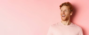 Kızıl saçlı ve sakallı yakışıklı beyaz adamın yakın çekimi, mutlu bir gülümsemeyle sola bakıyor, tanıtım teklifini kontrol ediyor, pembe arka planda duruyor..