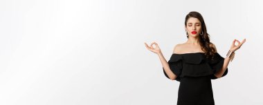 Sakin ve sabırlı genç esmer kadın meditasyon yapıyor, elleri zen jestli siyah elbiseli, beyaz arka planda duruyor..