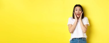 Güzellik ve moda konsepti. Şaşırmış Asyalı kız kameraya bakıp hayret ediyor. Reklamlara bakıyor, sarı arka planda duruyor..