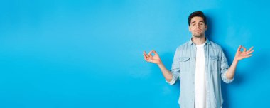 Sakin bir adam, gözleri kapalı meditasyon yapıyor, el ele tutuşuyor ve yoga yapıyor, mavi arka planda ayakta duruyor..